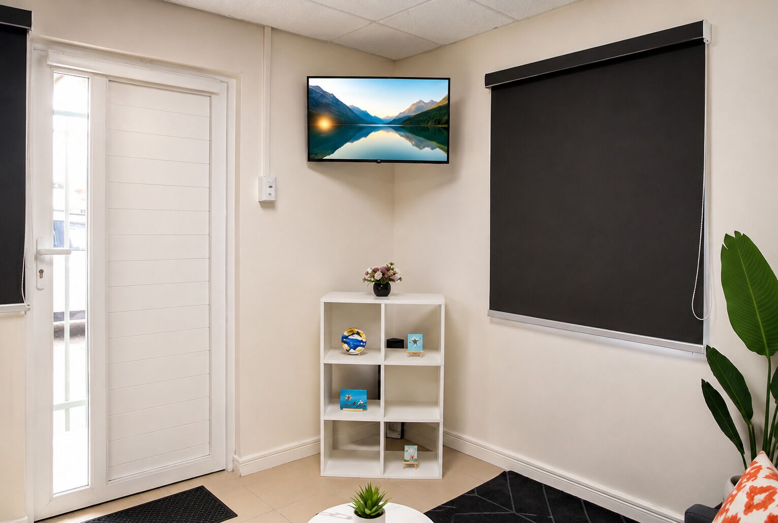 A room with a door, a white shelf with items, a TV on the wall, and a large window.
