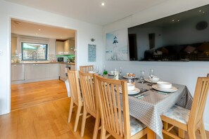 A dining area with a wooden table set for four, surrounded by chairs, in a modern kitchen with white cabinets and a large window. - 4 Bed in Westward Ho (oc-o45219) (Northam)