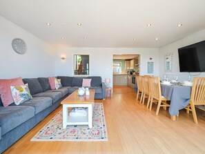 A modern living room with a grey sofa, a wooden coffee table, and a dining area with wooden chairs and a table set for dining. - 4 Bed in Westward Ho (oc-o45219) (Northam)