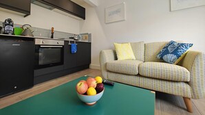 A modern kitchen with a built-in oven and a dining area with a green table and a bowl of fruit. - 1 Bed in Brixham (oc-f45535) (Brixham)
