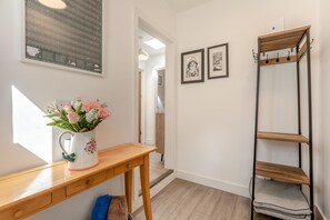 Ein Flur mit einem Holztisch, einer weißen Vase mit Blumen, einem Metallregal und zwei gerahmten Bildern an der Wand.