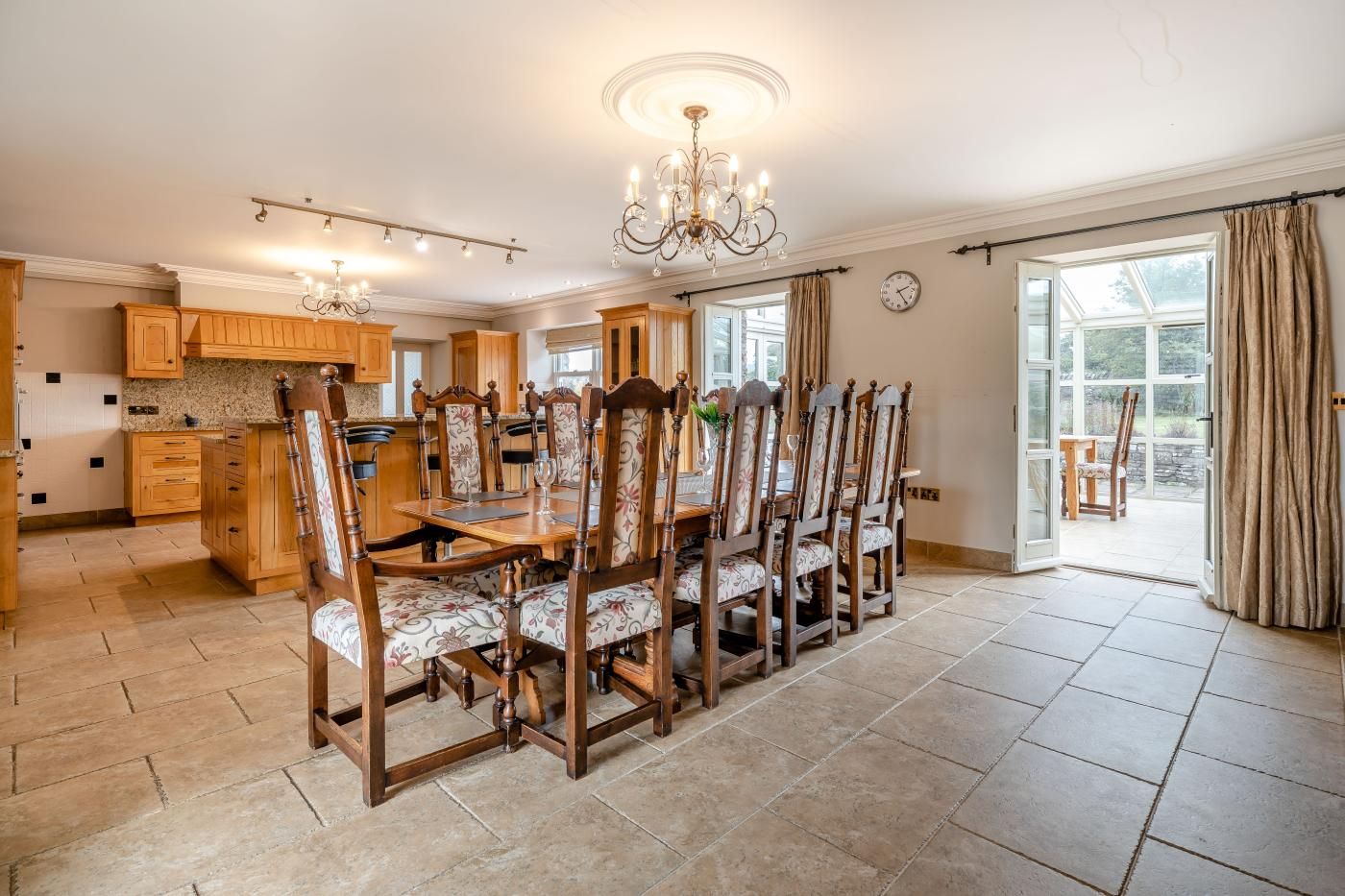 A spacious dining area with a long wooden table and chairs, a chandelier, and a view of the garden through large windows.
