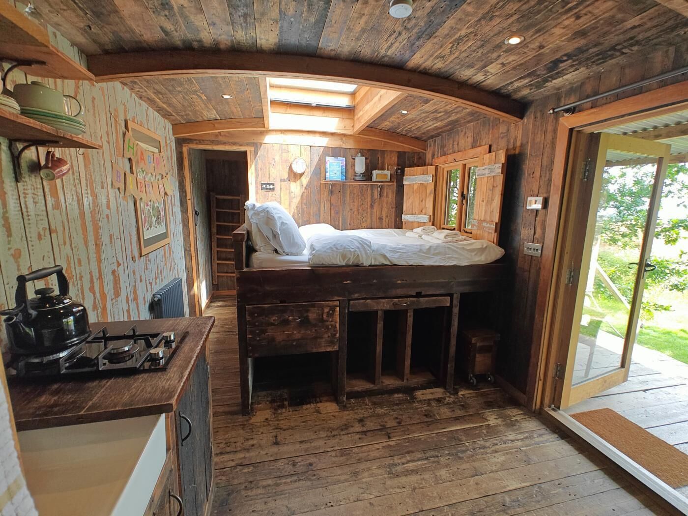 Interior rústico de una cabaña con literas, una pequeña cocina con estufa de gas y vistas al exterior a través de una puerta de cristal.