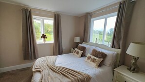 A bedroom with a large bed, two bedside tables, a window with curtains, and a view of greenery outside. - 3 Bed in Grizebeck (oc-b45345) (Kirkby Ireleth)