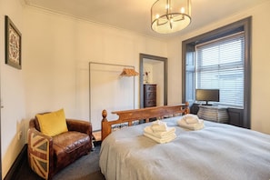 A bedroom with a large bed, a wooden headboard, a leather armchair, a bedside table with a lamp, and a window with blinds. - 3 Bed in Port William (oc-s45456) (Port William)