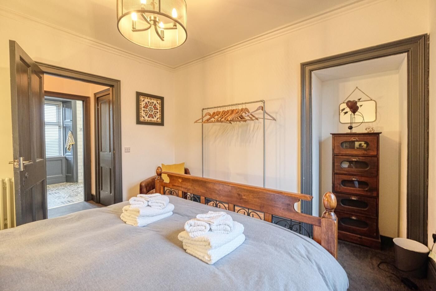 Una habitación de hotel con una cama grande, cabecera de madera, perchero, cómoda con cajones y un reloj de pared.