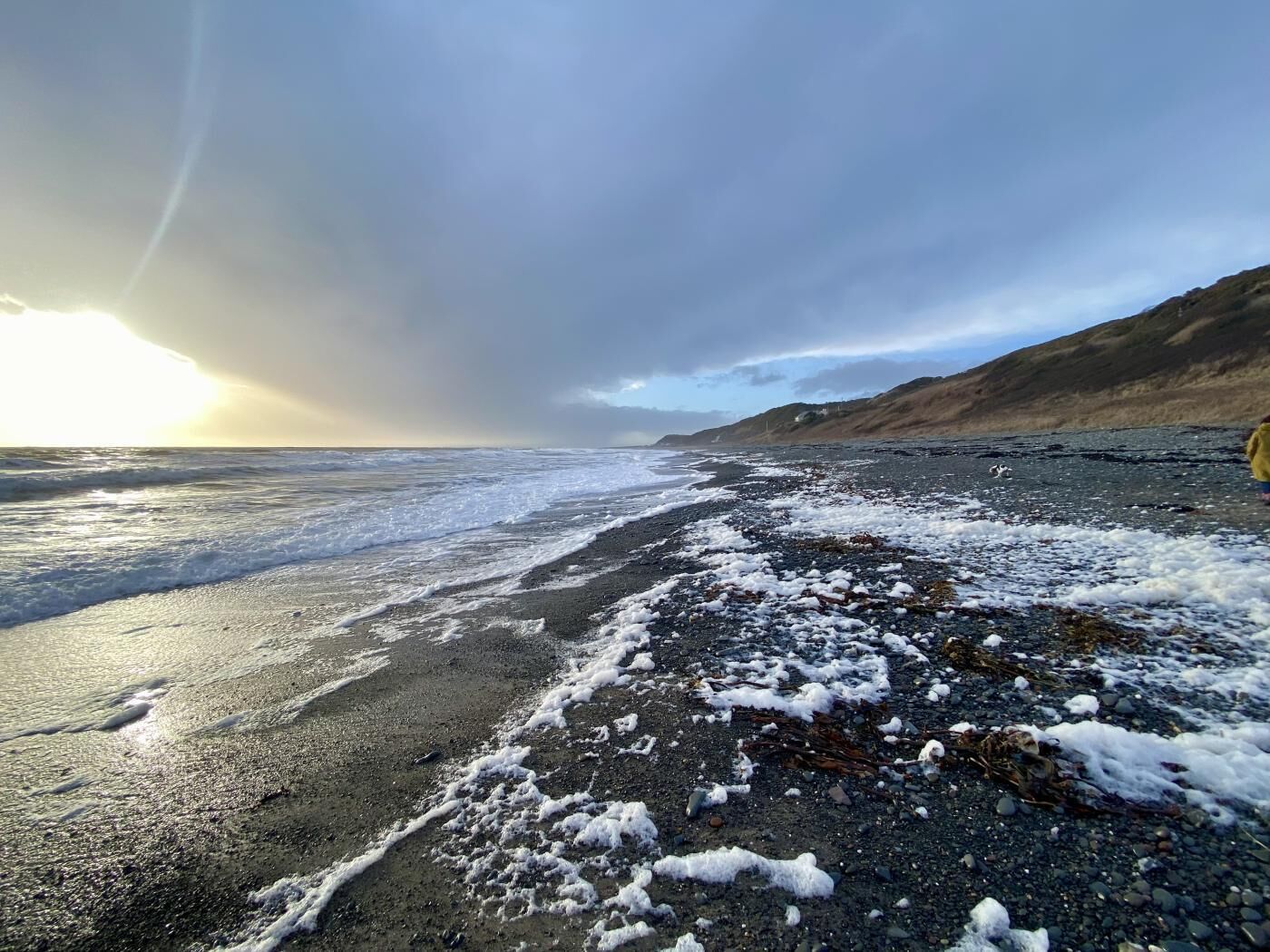 Een strand met golven, een bewolkte lucht en een verre vergezichten van heuvels.
