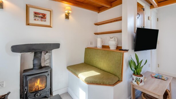 A cozy room with a wood-burning stove, a green cushioned bench, a flat-screen TV, and a small table with a plant. - 1 Bed in Ambleside (oc-a45864) (Ambleside)