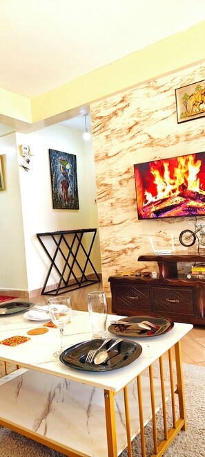 A dining area with a marble table, a TV displaying a fire, and a painting on the wall. - Modern 1BR Furnished Apartment with Balcony and Work-From-Home Space in Ruaka (Nairobi)