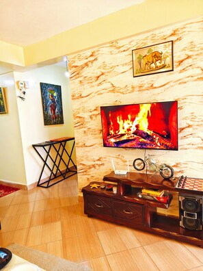 A living room with a flat-screen TV mounted on a marble wall, a wooden coffee table with decorative items, and a fireplace with a visible fire. - Modern 1BR Furnished Apartment with Balcony and Work-From-Home Space in Ruaka (Nairobi)