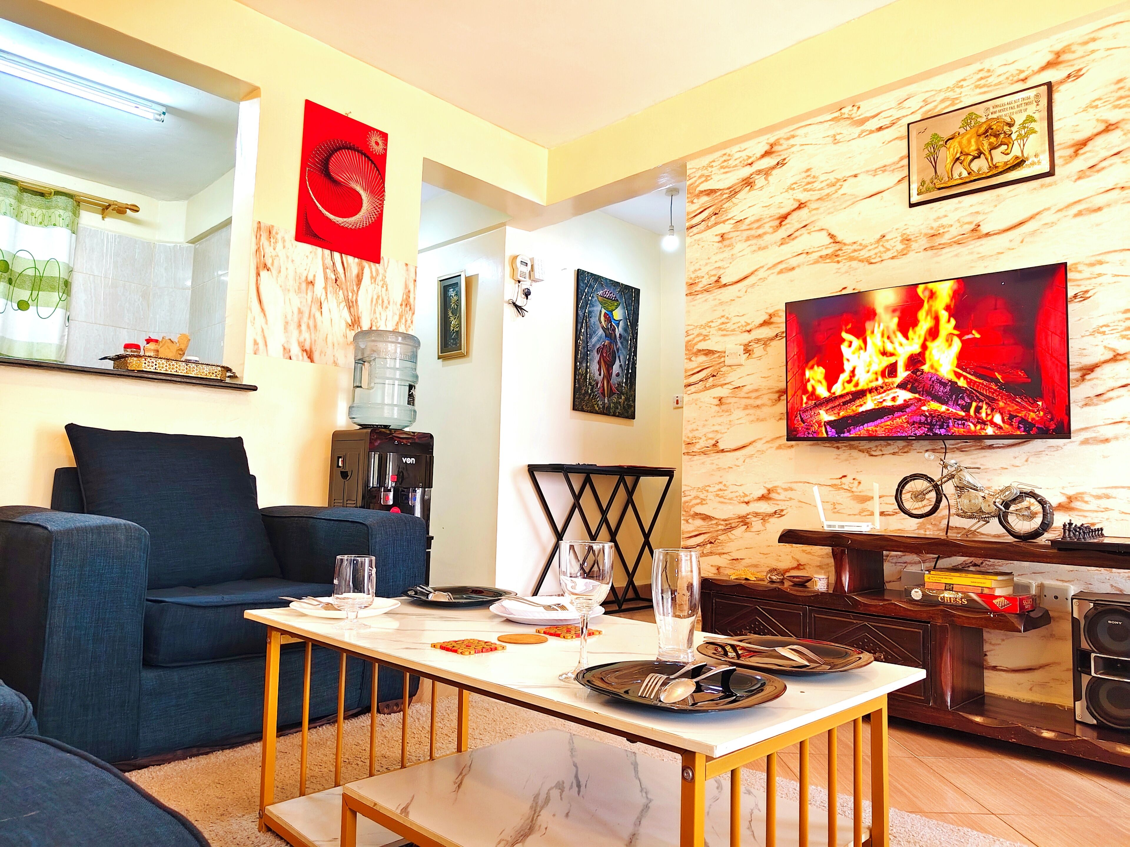 A living room with a sofa, a coffee table, and a TV displaying a fire. There is a water dispenser and a framed picture on the wall.