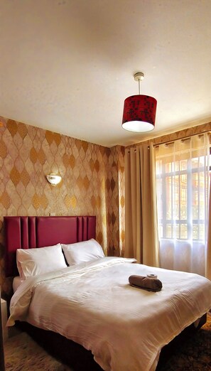 A hotel room with a bed, a headboard, a wall-mounted light, and a window with curtains. - Modern 1BR Furnished Apartment with Balcony and Work-From-Home Space in Ruaka (Nairobi)