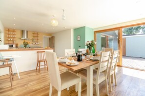 A dining area with a wooden table, white chairs, and a kitchen in the background. - 4 Bed in Beccles (oc-b45848) (Beccles)