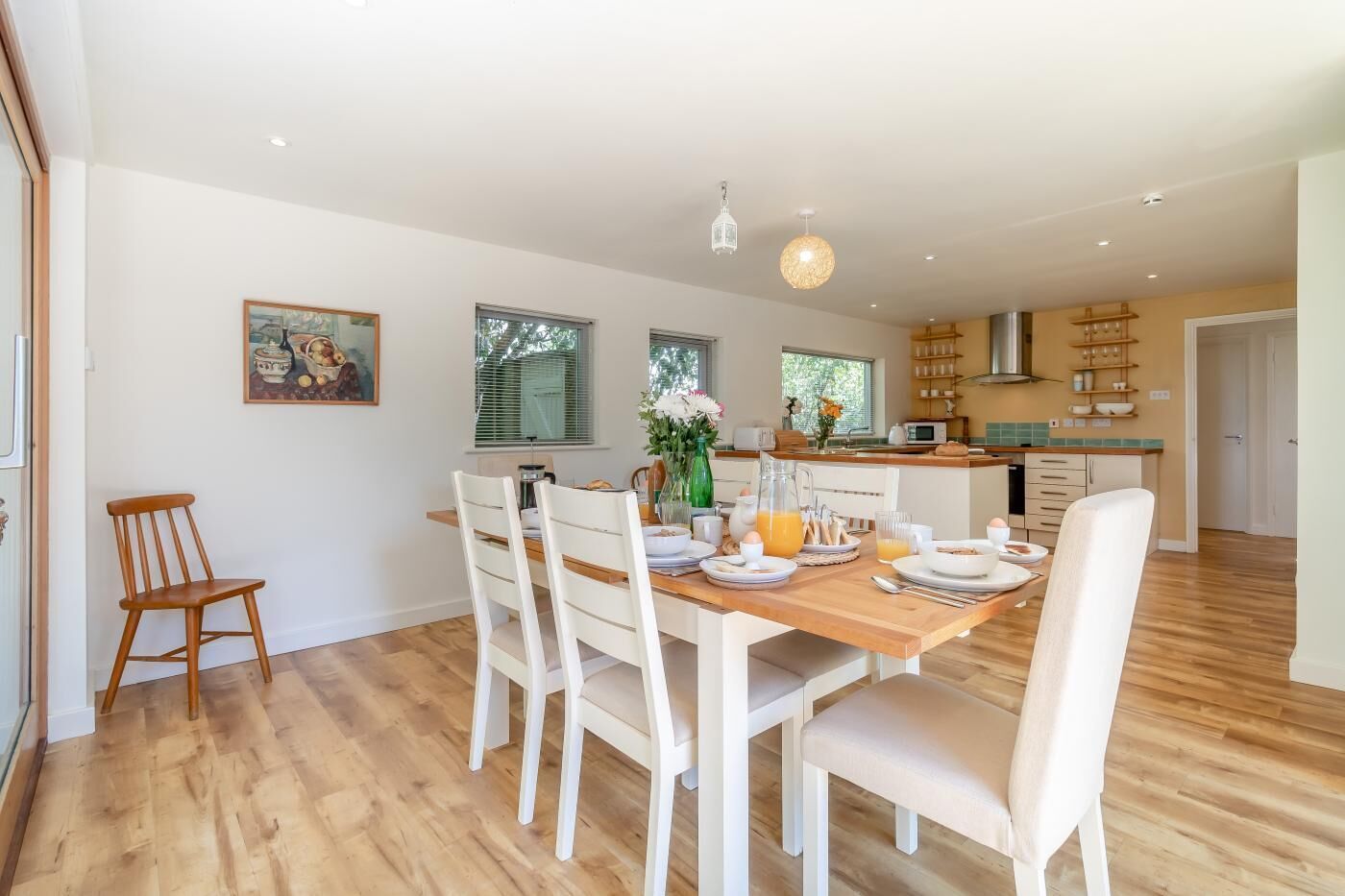 A dining area with a wooden table set for breakfast, white chairs, and a kitchen in the background.