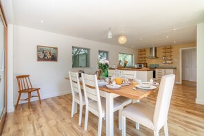 A dining area with a wooden table set for breakfast, white chairs, and a kitchen in the background. - 4 Bed in Beccles (oc-b45848) (Beccles)
