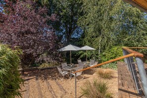 An outdoor patio with a table and chairs, surrounded by trees and a wooden deck. - 4 Bed in Beccles (oc-b45848) (Beccles)