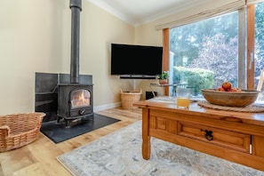 A living room with a wood-burning stove, a television mounted on the wall, a dining table with a bowl of fruit, and a view of greenery outside. - 4 Bed in Beccles (oc-b45848) (Beccles)