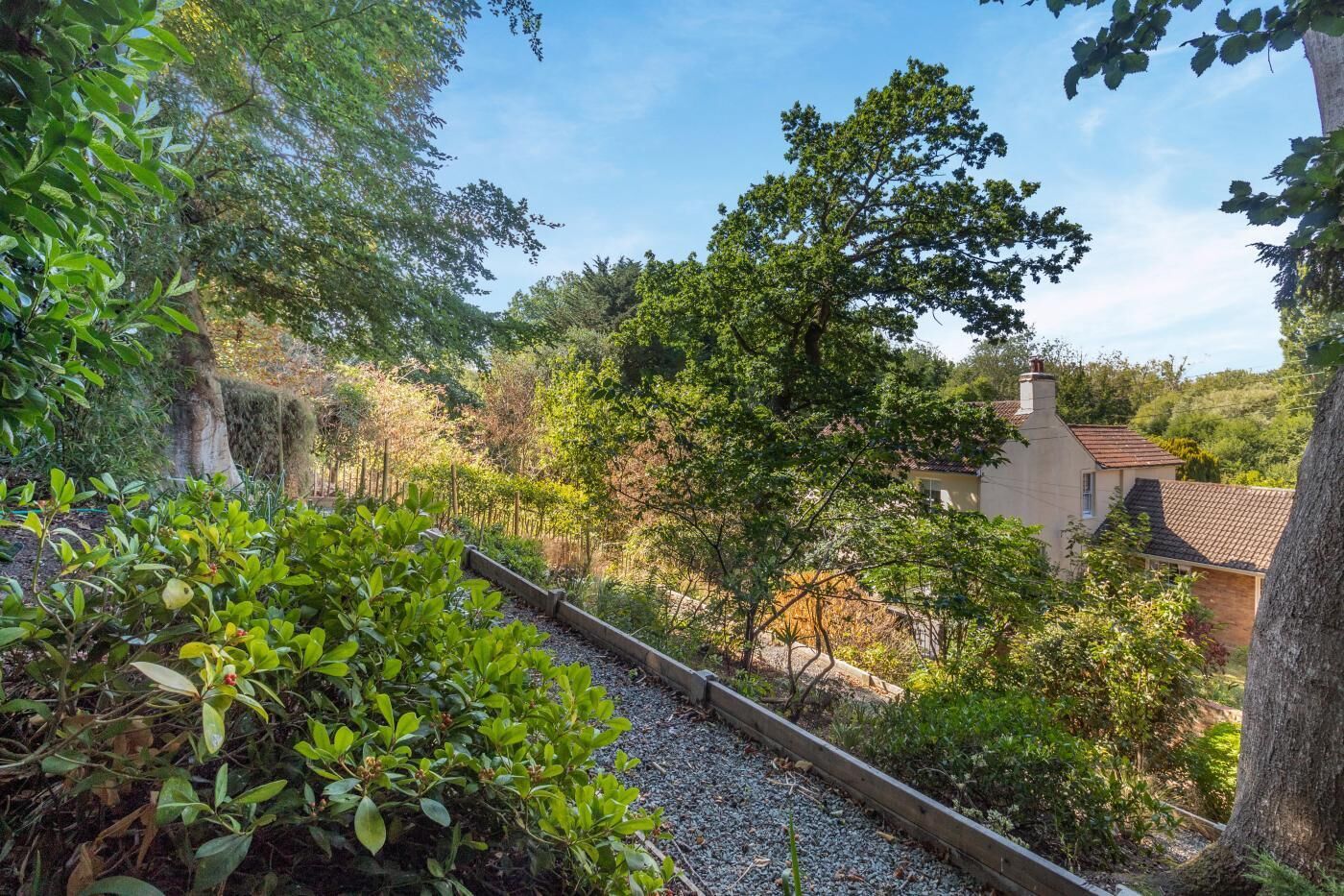 Un jardin avec un chemin de gravier, un feuillage dense et un bâtiment doté d’un toit en tuiles.