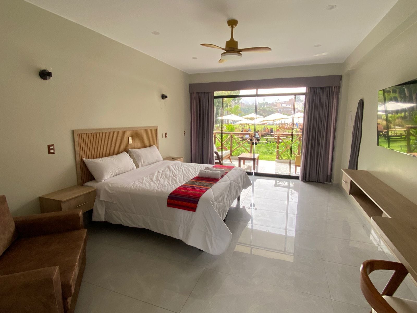 Habitación de hotel con una cama grande, ventilador de techo, televisor y balcón con mobiliario exterior.
