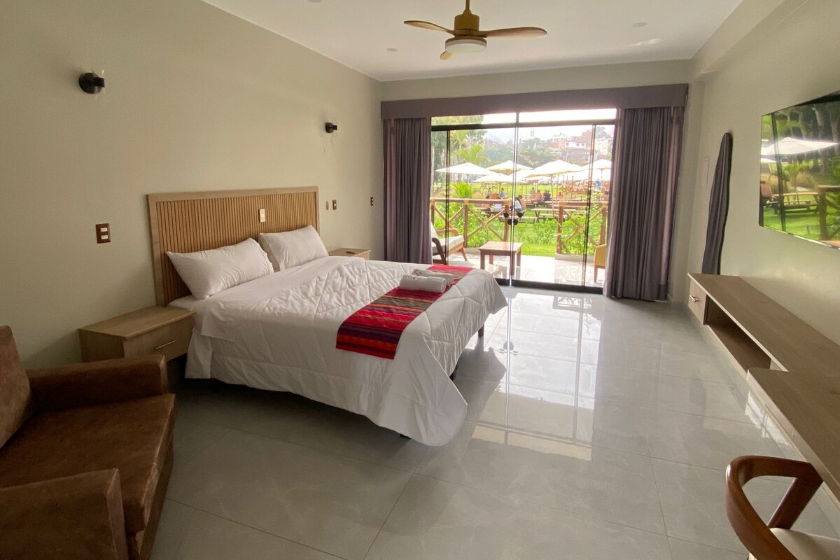 A hotel room with a large bed, a ceiling fan, a TV, and a balcony with outdoor seating.