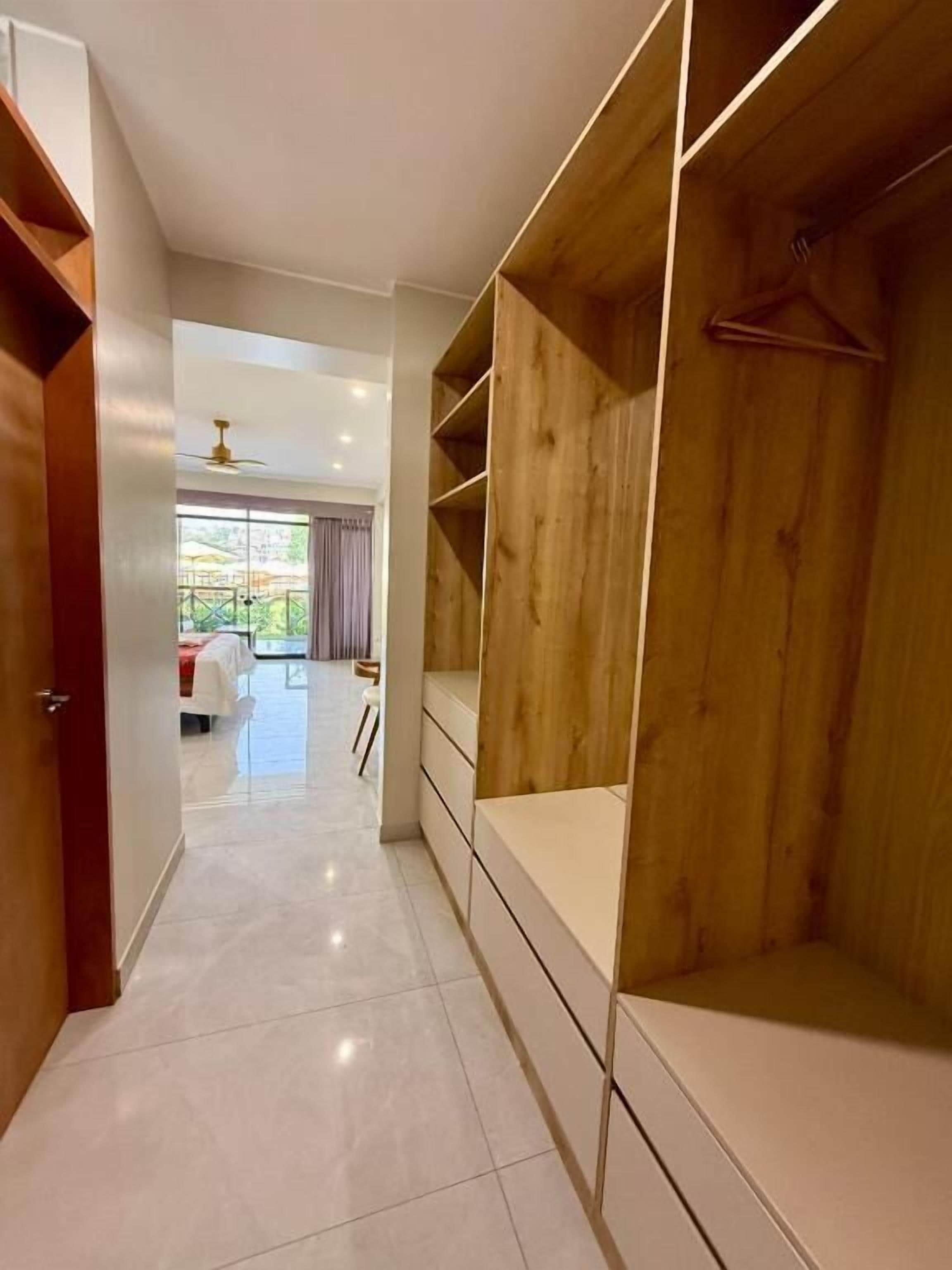 Een gang met een houten garderobe, een kledingroede en een zicht op een kamer met een bed en gordijnen.