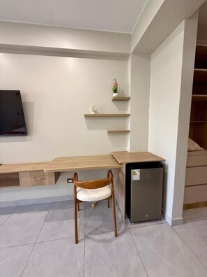 A compact room with a wooden desk, a chair, a small refrigerator, and a TV mounted on the wall.