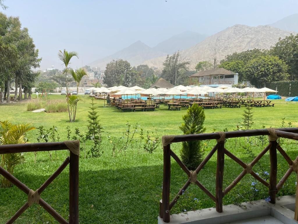 Un espace extérieur verdoyant avec des tables de pique-nique et des parasols, une clôture en bois et des montagnes en arrière-plan.