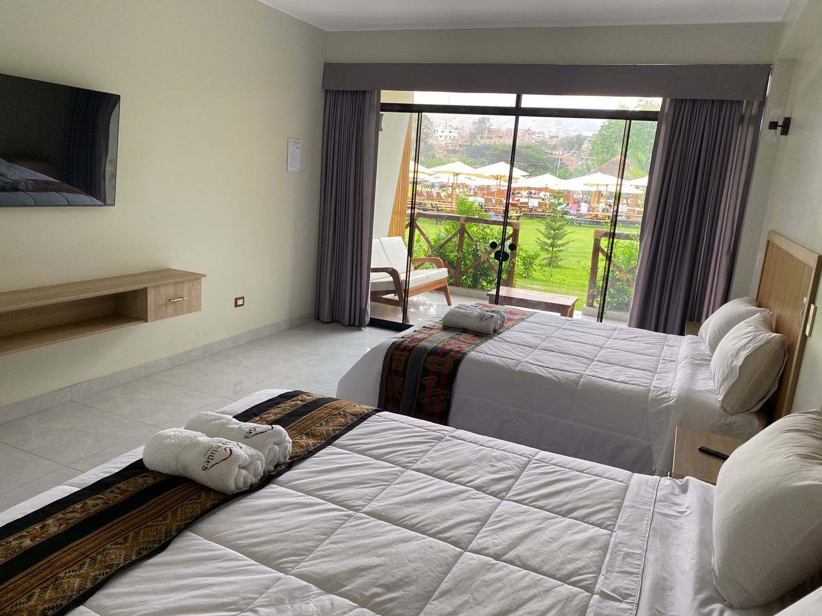 Habitación de hotel con dos camas, un televisor montado en la pared y un balcón con vistas a la ciudad.