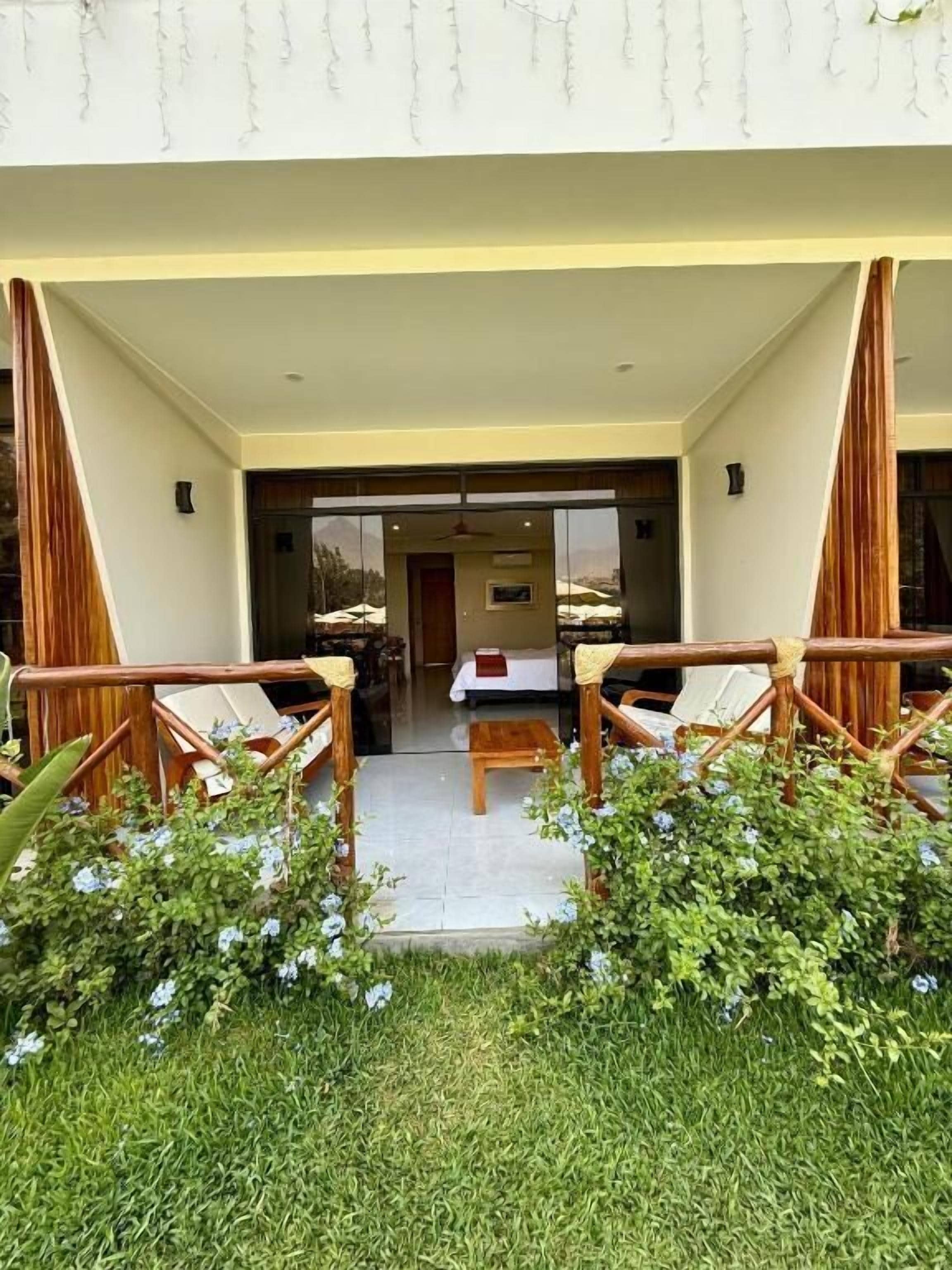 Un espacio de estar al aire libre con techo, con dos sillas de madera y una mesa, rodeado de follaje y vistas al interior.