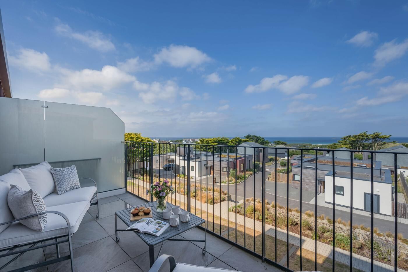 Un balcon avec un canapé blanc, une petite table sur laquelle se trouve un livre et un vase de fleurs, et une vue sur un quartier résidentiel et l’océan.