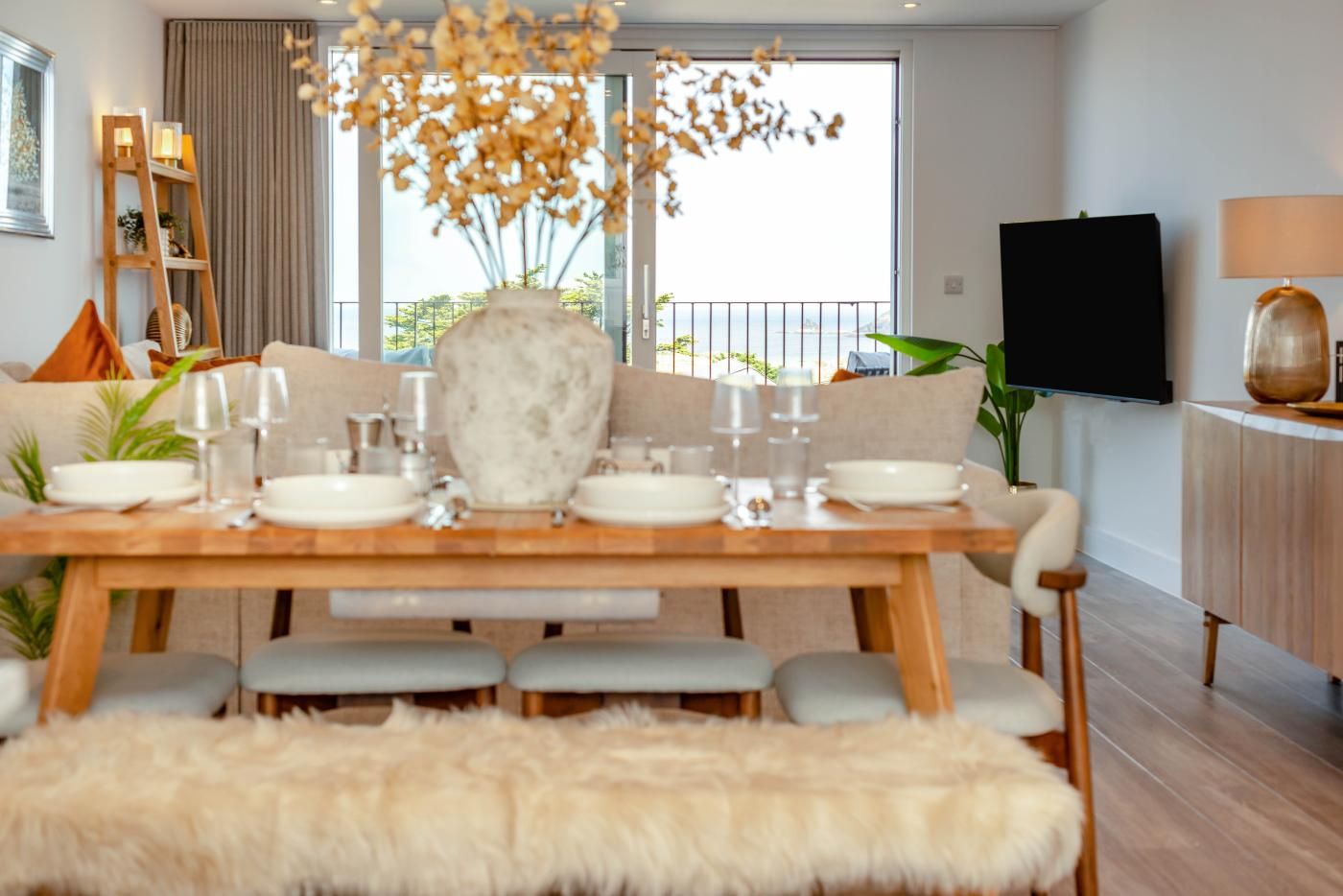 Ein Essbereich mit einem Holztisch, gedeckt für vier Personen, einer großen Vase mit Trockenblumen und einer Couch mit Kissen.