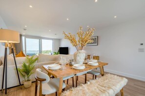 Ein moderner Essbereich mit einem Holztisch, gedeckt für vier Personen, einer Vase mit gelben Blumen und einer Couch im Hintergrund.