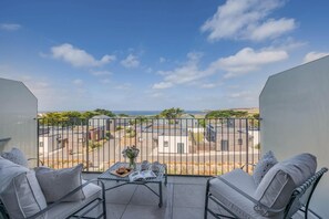 Un balcon avec deux chaises, une petite table sur laquelle se trouvent un livre et une assiette de nourriture, et une vue sur un quartier résidentiel avec des maisons et un ciel dégagé.