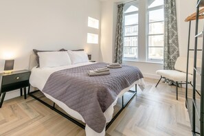 Ein ordentlich bezogenes Bett mit gesteppter Decke, ein Nachttisch mit Lampe und ein Sessel an einem fenster mit Vorhängen.