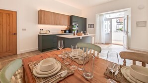 Uma cozinha moderna com armários de madeira, um espaço de jantar com uma mesa de madeira posta para quatro pessoas e vista para o exterior por uma porta aberta.