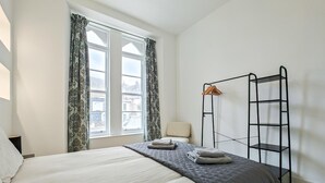 Quarto com cama, janela com cortinas, uma cadeira e estante de metal.