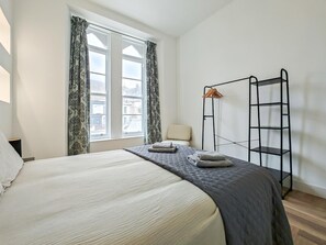 Ein Schlafzimmer mit einem Bett, einem Fenster mit Vorhängen, einem Sessel und einem Metallregal.