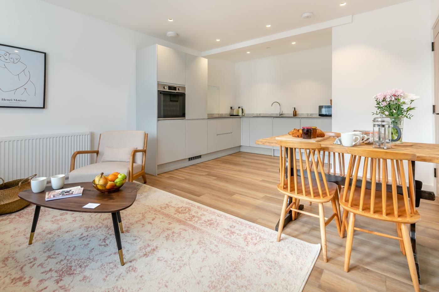 Une cuisine moderne avec des armoires blanches, un four encastré et un coin repas comprenant une table et des chaises en bois.