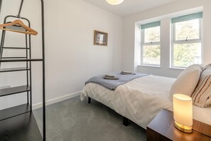 A bedroom with a bed, a bedside table with a lamp, a window with a view, a wall-mounted shelf, and a picture frame. - 2 Bed in Ilfracombe (oc-b45844) (Ilfracombe)