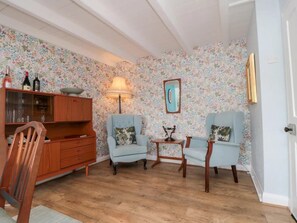 Ein Zimmer mit floraler Tapete, einem Holzschrank, zwei Sesseln, einem kleinen Tisch und einer Lampe.
