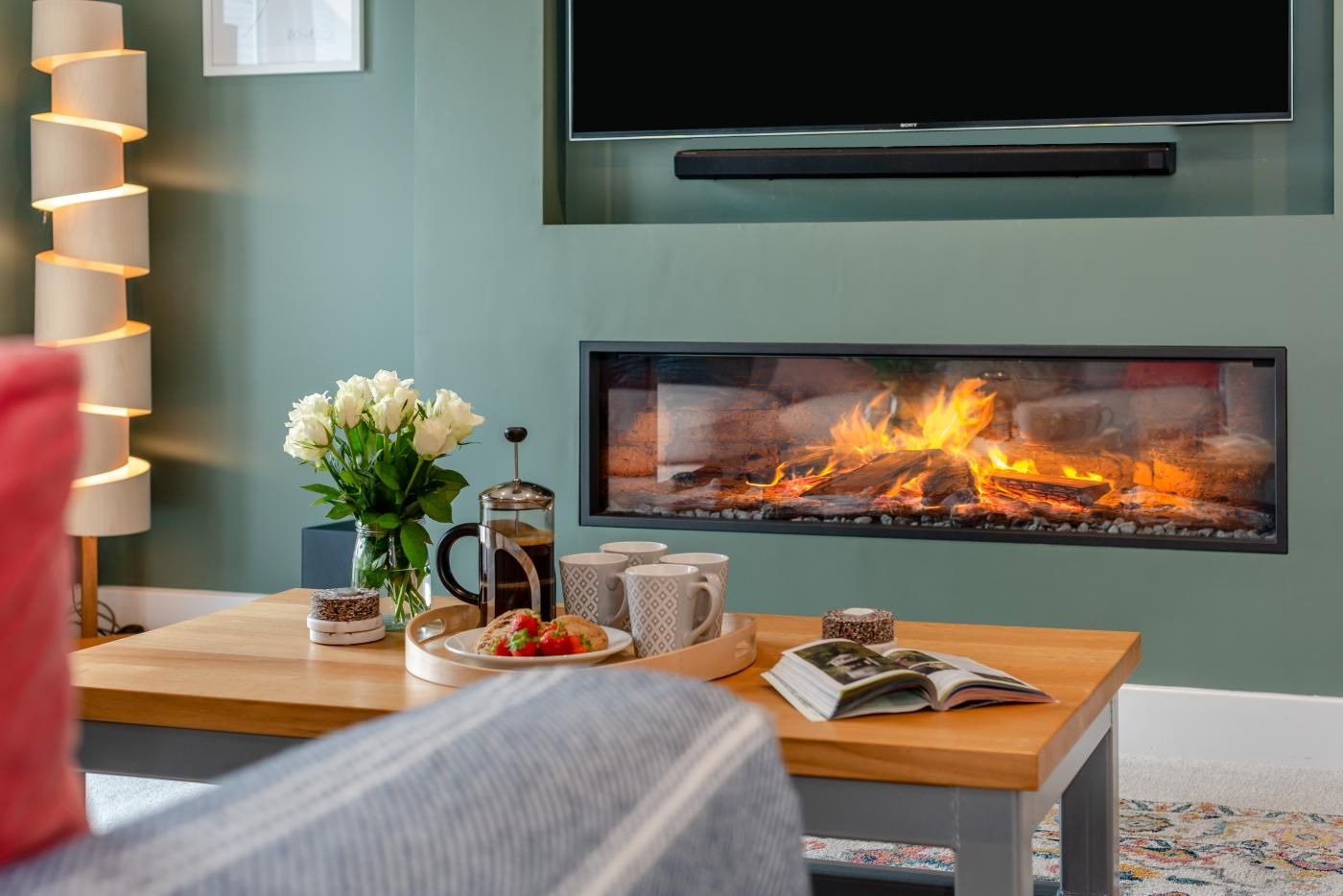 A living room with a fireplace, a coffee table with a book, a vase of flowers, and a television mounted on the wall.