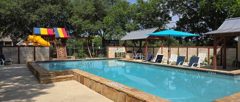 A swimming pool with a slide, lounge chairs, and a covered area with umbrellas.