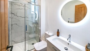 Ein modernes Badezimmer mit einer Duschkabine aus Glas, einem runden Spiegel und einem weißen Waschbecken mit Holztür.