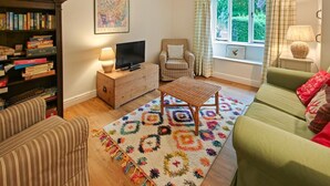 Ein Wohnzimmer mit einer Couch, einem Fernseher auf einem Holzschrank, einem Bücherregal, einem gemusterten Teppich und einem Fenster mit Vorhängen.