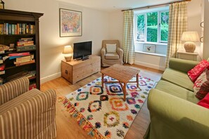 Ein Wohnzimmer mit einer Couch, einem Fernseher auf einem Holzschrank, einem Bücherregal, einem gemusterten Teppich und einem Fenster mit Vorhängen.