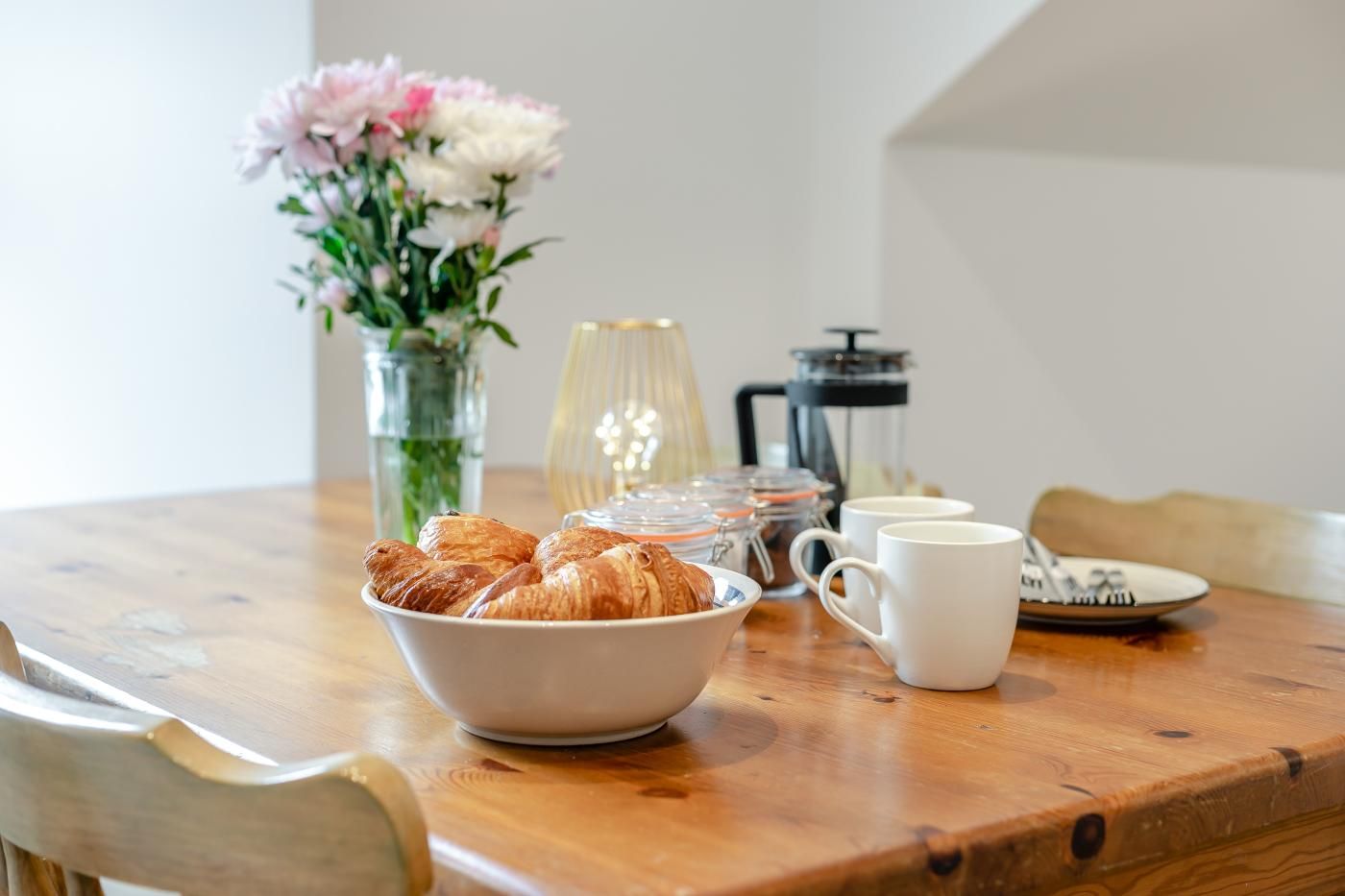 Une table en bois sur laquelle se trouvent un panier de croissants, une cafetière et un vase de fleurs.