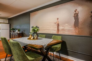 A dining area with a table set for four, surrounded by green upholstered chairs. A large painting of a serene lake scene is on the wall behind the table. - Cozy 2-bedroom in Cotter with walking distance to shopping, river, and food. (Cotter)