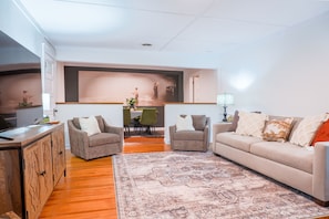 A living room with a sofa, two armchairs, a coffee table, a TV, a dining table with chairs, and a patterned rug. - Cozy 2-bedroom house in Cotter, walking distance to shopping, river, and food. (Cotter)