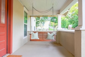 A porch with a swing, a red door, and a view of a house and trees. - Cozy 2-bedroom house in Cotter, walking distance to shopping, river, and food. (Cotter)
