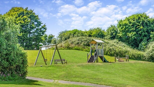 En grøn park med gyngestativ, et trælegetårn og en bænk.